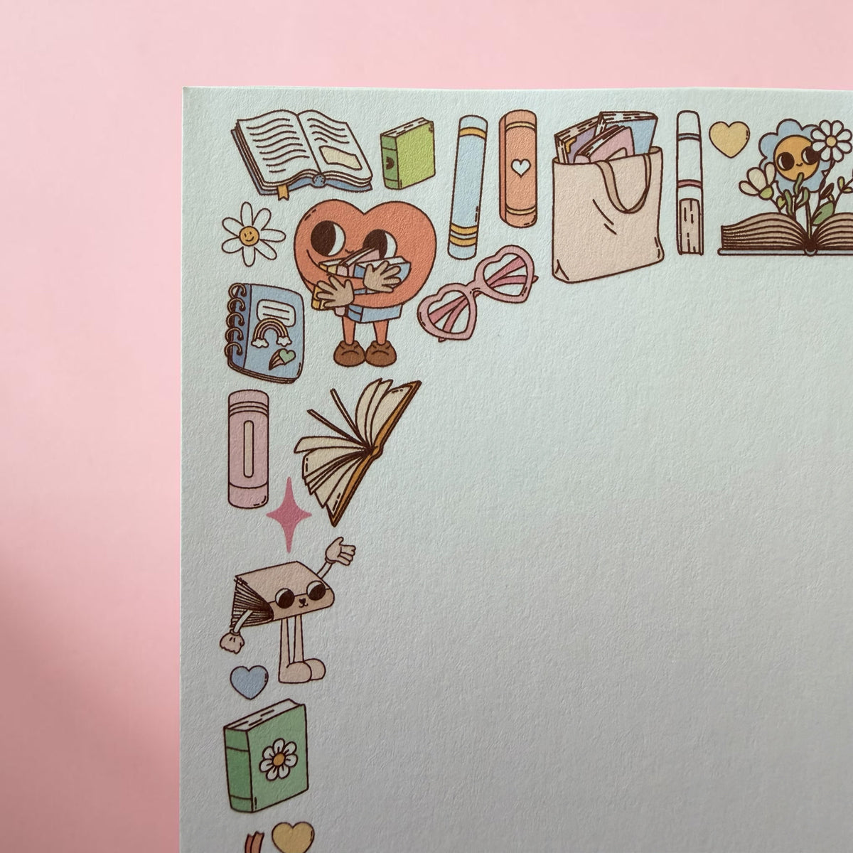 A close-up of a pastel stationery sheet or card with cute illustrated school and study-themed icons along the top and left edges, including books, a tote bag, glasses, flowers, and small doodles, set against a soft pink background.