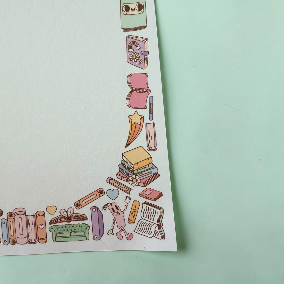 A close-up of a white sheet of paper with a decorative border of cute, whimsical book-themed illustrations in pastel colors, placed on a mint green background.