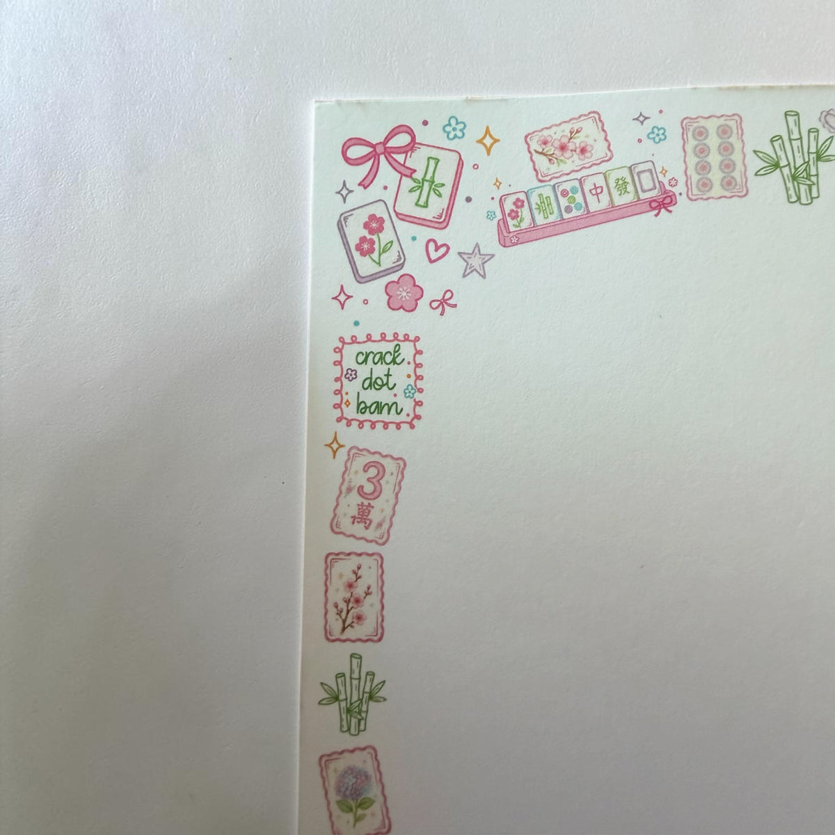 A close-up of a white paper corner decorated with pink and green Japanese mahjong-themed washi tape stickers, including tiles, bamboo, stars, and the text 'crack dot bam'.