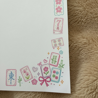 A close-up of a white sheet of paper printed with colorful mahjong-themed illustrations along the bottom and right edges, placed on a beige textured fabric surface.
