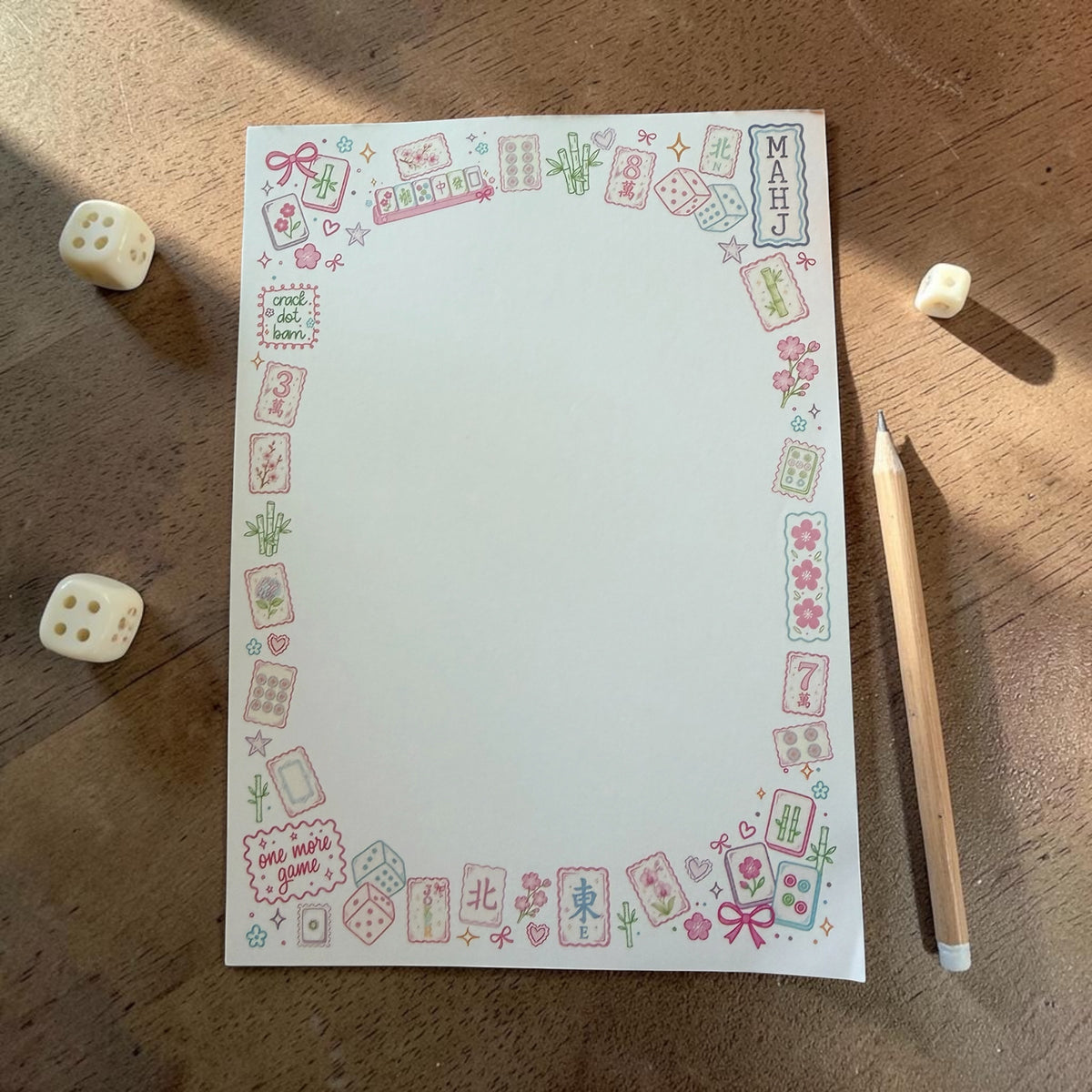 A square white notepad with a colorful Japanese mahjong-themed border, displayed on a wooden tabletop next to dice and a pencil.
