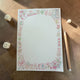 A square white notepad with a colorful Japanese mahjong-themed border, displayed on a wooden tabletop next to dice and a pencil.